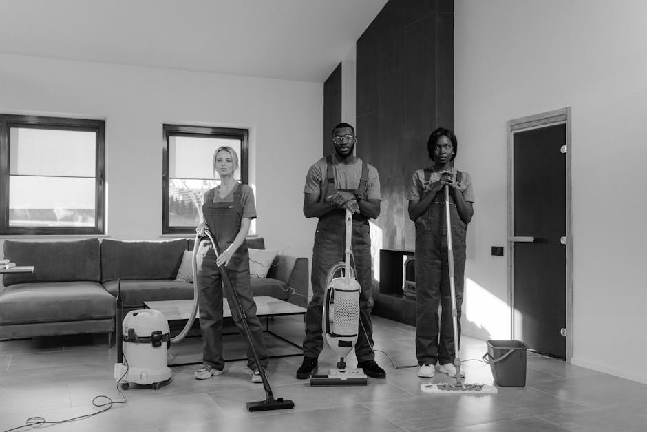 A team of three professional cleaners working in a modern living room with a minimalist design, featuring a large sectional sofa and two windows allowing natural light to fill the space. The cleaners are dressed in uniform overalls; one on the left is using a vacuum cleaner on the light-colored wooden floor, while the one in the center is holding a vacuum attachment and standing confidently. The third cleaner on the right is pushing a mop with a bucket nearby, preparing to clean the tiled area near a plain door. The room's surfaces, including the wooden flooring and tiled sections, appear to be in the process of thorough cleaning, highlighting attention to hygiene and surface maintenance. Cleaner Harrow offers domestic cleaning services focused on surface cleaning and sanitisation, as demonstrated by the team's equipment and workflow in this setting.