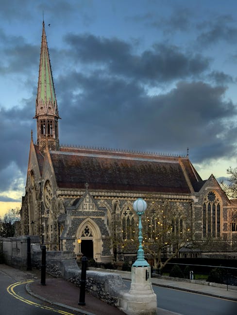 An exterior view of a historic stone church with intricate Gothic architecture, tall pointed spire, stained glass windows, and detailed stone carvings. The church is illuminated by street lamps, with a cloudy evening sky overhead, creating a dramatic backdrop. The surrounding area features a paved pathway, bollards, and leafless trees, suggesting an urban setting near Harrow on the Hill. The image emphasizes the church's weathered stone surface and well-maintained appearance, which may benefit from professional cleaning services offered by Cleaner Harrow, especially for deep cleaning and surface maintenance of historic structures.