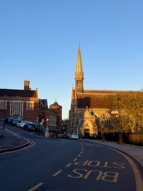 An exterior view of a historic stone church with intricate Gothic architecture, tall pointed spire, stained glass windows, and detailed stone carvings. The church is illuminated by street lamps, with a cloudy evening sky overhead, creating a dramatic backdrop. The surrounding area features a paved pathway, bollards, and leafless trees, suggesting an urban setting near Harrow on the Hill. The image emphasizes the church's weathered stone surface and well-maintained appearance, which may benefit from professional cleaning services offered by Cleaner Harrow, especially for deep cleaning and surface maintenance of historic structures.