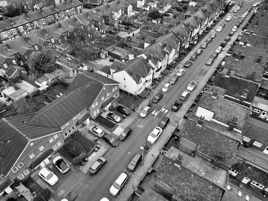 An aerial black-and-white photograph of a residential neighborhood in Wealdstone and Headstone Lane Harrow, showing rows of terraced houses and detached homes. The houses have pitched tiled roofs, some with dormer windows, and small front gardens. A narrow street runs through the center, lined with parked cars on both sides, including sedans and hatchbacks, with a few cars in motion. Sidewalks run along the street, with streetlamps and road markings visible. Gardens and backyards are seen behind some homes, with greenery and fencing. The overall scene depicts a quiet, well-organized suburban area with dense housing and consistent street layout.