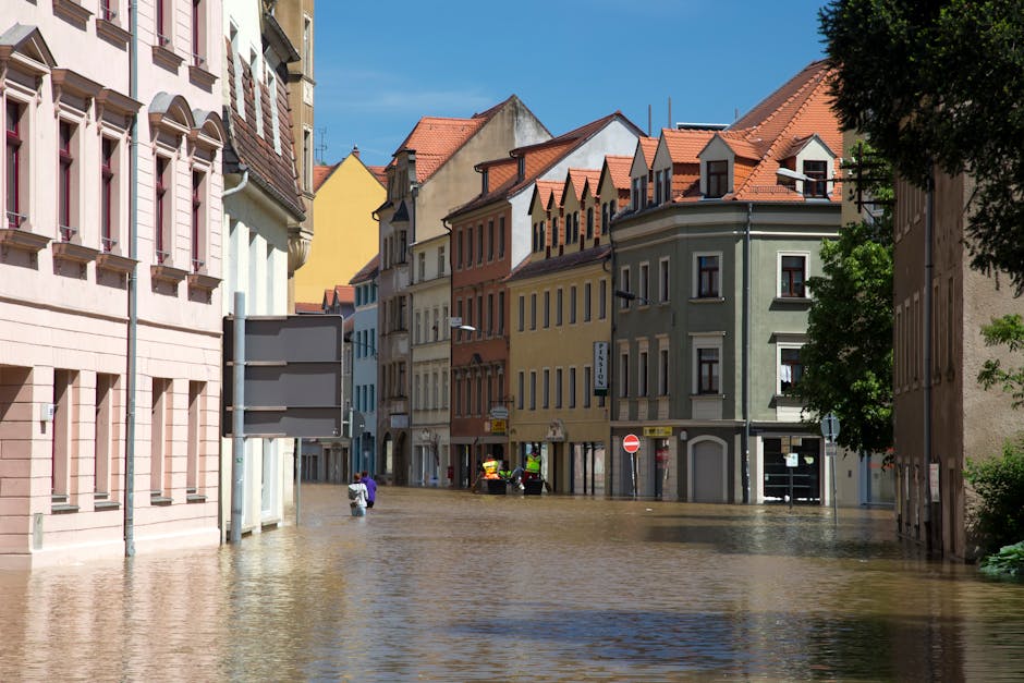 A flooded street in a residential area featuring multi-story buildings with pastel-colored facades, including pink, yellow, and green, along with red-tiled roofs. The water has risen to cover the cobblestone pavement, reaching the windows and doors, with some vehicles and pedestrians partially submerged. Bright sunlight illuminates the scene, highlighting the wet surfaces and casting shadows from nearby trees and building structures. This image reflects the aftermath of a flood in an urban environment, emphasizing the importance of surface cleaning and maintenance to restore hygiene and safety, as provided by Cleaner Harrow. The scene is devoid of visible cleaning tools or personnel, focusing solely on the flood impact on the residential street.