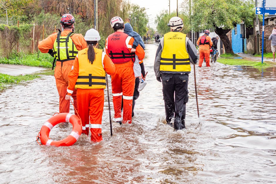 A group of emergency responders wearing bright orange and yellow waterproof jackets and helmets are wading through a flooded street, with some holding rescue poles and a life ring. The street is submerged in muddy water, reaching halfway up their boots, and is lined with trees and small buildings. Overhead, the sky is overcast, and in the background, other rescuers and residents are seen managing the flood situation. The scene emphasizes the efforts of emergency services during flood incidents, with visible water covering the surface of the road and creating a challenging environment. This image relates to emergency flood response, similar to the swift flood cleaning services provided by Cleaner Harrow, who specialize in domestic and commercial property sanitation after flooding incidents.