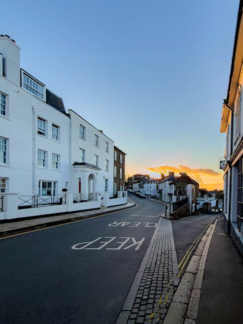 A residential street scene taken during sunset, featuring clean, well-maintained white and brick buildings with modern design, large windows, and balconies. The asphalt road has clear white markings, including a 'KEEP CLEAR' notice, and the pavement is composed of small, interlocking bricks. The street is illuminated by natural light from the setting sun, casting warm hues across the scene, with no visible litter or debris, indicating regular cleaning and maintenance. This image represents an urban environment in Harrow, suitable for illustrating a professional domestic or commercial cleaning service such as that provided by Cleaner Harrow at the end of tenancy or surface cleaning for flats on Station Road.