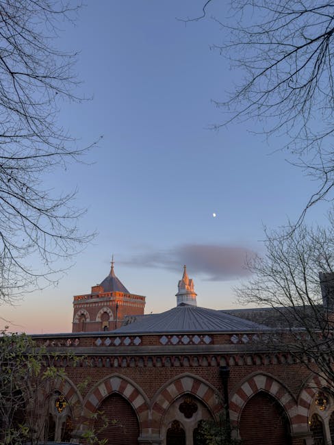 The image depicts a clear evening sky over a historic brick building with distinctive red and white arches, domes, and towers. Bare tree branches frame the scene, and a small bright moon is visible above, casting a serene atmosphere. The lighting suggests the scene is captured at dusk, with soft natural light highlighting the architectural details. This setting emphasizes the clean and well-maintained appearance of the exterior structure, which aligns with the cleaning, sanitisation, and maintenance services provided by Cleaner Harrow, especially relevant to end-of-tenancy cleaning for flats on Station Road Harrow in the HA1 postcode area.