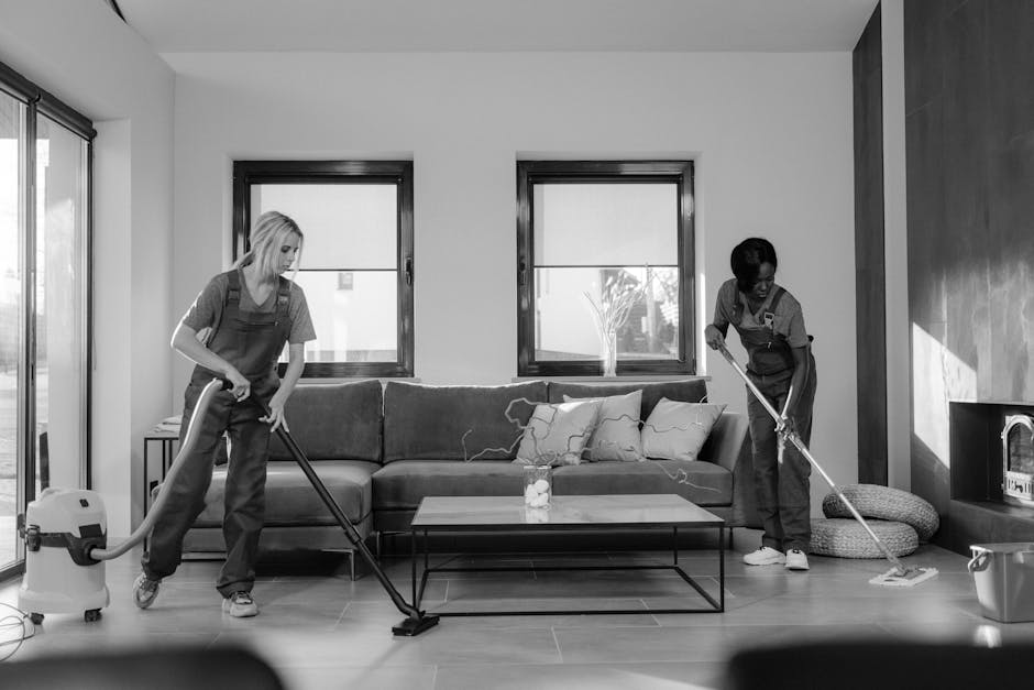 Three professional cleaners, two women and one man, dressed in matching grey t-shirts and bright orange overalls, are standing in a modern living room with white walls, a grey sofa, and large windows.